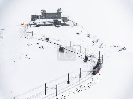 Aerial shot shows a red and white Swiss train climbing Zermatts snowy slopes, Switzerland. Tracks cut through snow, revealing rocks and a dome building against a cloudy sky.