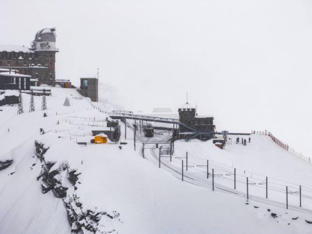 Eine heitere, verschneite Szene in Zermatt in den Schweizer Alpen zeigt einen gelben Zug in den Bergen. Schnee bedeckt das Gebiet, eine Mischung aus traditioneller und moderner Architektur, mit Skifahrern für Skala.