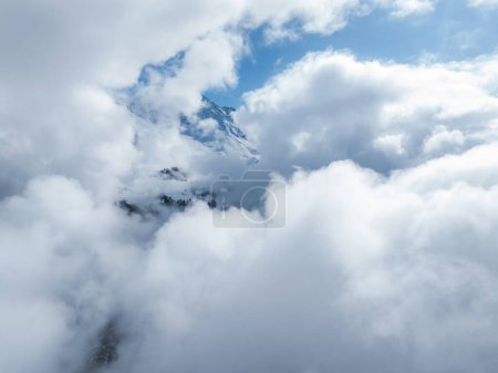 A breathtaking aerial view of the Swiss Alps near Verbier, with majestic peaks peeking through a dense cloud blanket. Snow covered slopes and rugged terrain contrast with a serene blue sky.