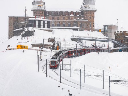 Aerial view of a red train ascending a snow covered track at Zermatt ski resort, with a historic stone building in the background. Overcast skies and a winter atmosphere highlight the scene.