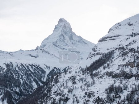 Iconic Matterhorn in Zermatt, Switzerland, shows its pyramid peak against a pale sky. Early or late light highlights its snow covered slopes and dark forests.