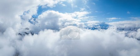 An aerial view over Verbier, Switzerland, reveals dense clouds and mountain peaks. The vivid blue sky contrasts white clouds, showcasing the Swiss Alps serene beauty.