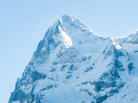 A majestic, snow covered mountain peak in the Swiss Alps near Murren basks in soft sunlight. Its rugged slopes, sharp ridges, and crevasses are highlighted against a serene, pale blue sky.