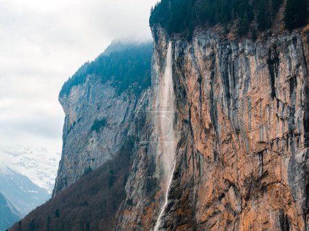 A stunning waterfall flows down cliffs in Murren, Switzerland, amid a misty, mountainous scene. Its geological layers and lack of human buildings emphasize its natural beauty.