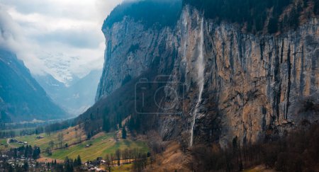 Photo for Aerial shot of Murren, Switzerland shows its cliffside spot in a lush valley. It features farmlands, a rocky cliff, and the Swiss Alps under soft, overcast light for a dramatic, moody scene. - Royalty Free Image
