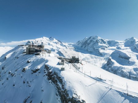 A stunning aerial shot of Zermatt, Switzerlands snowy mountains, with a train on a narrow track and a ski lift. The clear blue sky and deep snow showcase its winter sports allure.