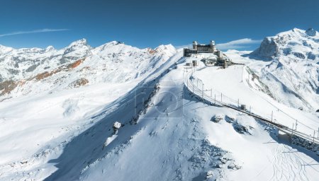 Eine Luftaufnahme zeigt Zermatt in der Schweiz mit einer verschneiten Landschaft und einem Bahngleis zu einem Gipfel bei klarem Himmel. Zerklüftete Berge im Hintergrund unterstreichen die heitere Schönheit der Höhenlage.