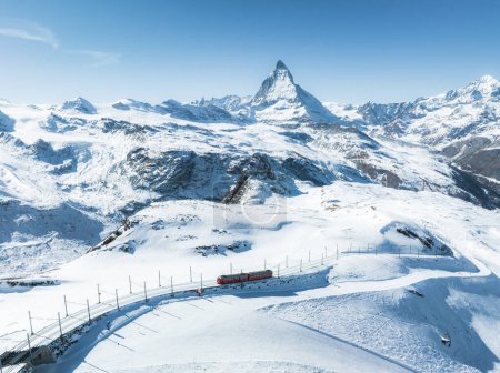 A stunning aerial shot of Zermatt ski resort, featuring a red train crossing snowy terrain with the Matterhorn peak and a clear blue sky in the backdrop. It showcases the Swiss Alps winter beauty.