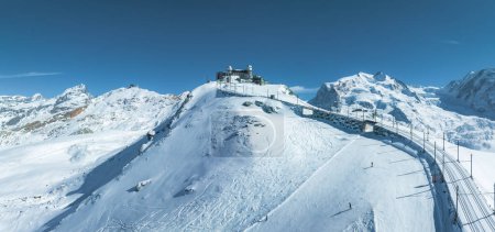 Eine Luftaufnahme des Skigebiets Zermatt in der Schweiz zeigt einen Zug, der verschneite Hänge, eine Gipfelanlage und Skifahrer auf dichtem Schnee erklimmt. Klarer Himmel und alpine Gipfel bieten eine atemberaubende Kulisse.