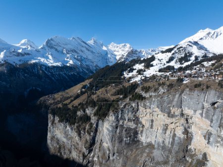 Foto de Una vista aérea de Murren, Suiza, revela chalets cubiertos de nieve en una pendiente y edificios cerca de un acantilado que conduce a una garganta, todo contra picos nevados y un cielo azul claro. - Imagen libre de derechos