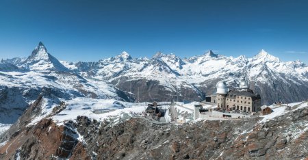 An aerial view of Zermatt ski resort in the Swiss Alps shows the Matterhorn, snow peaks, a train station, and a stone observatory, blending nature with human structures.