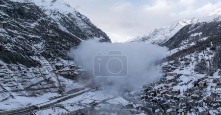 Téléchargez les photos : Vue aérienne de la station de ski de Zermatt, Alpes suisses, montre des montagnes enneigées, la ville dans la vallée, et une pente d'escalade de train. Nuages denses et lumière précoce ou tardive rehaussent la scène sereine. - en image libre de droit