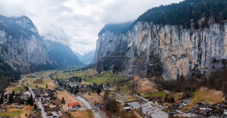 Aerial shot of Murren, Switzerland, shows alpine buildings and a road by a winding river. Cliffs and misty mountains surround the serene valley town under a cloudy sky, emphasizing its rugged charm.