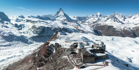 A stunning aerial shot of Zermatt ski resort shows a train climbing snow capped mountains, with the iconic Matterhorn in the backdrop. Buildings sit on the slope, beneath a clear blue sky.