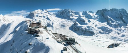 Aerial shot of Zermatt ski resort, Swiss Alps, with snow peaks and a train on a narrow railway up the mountain. Alpine buildings and winter sports facilities under a clear blue sky.