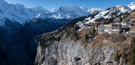 Aerial view of Murren, Switzerland, perched on a cliff with snow capped Swiss Alps in the background. Traditional alpine buildings dot the landscape, surrounded by forests under a clear blue sky.