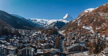 Aerial shot of Zermatt, Switzerland, displays chalets and hotels with snow roofs. The Matterhorn peak stands out, with a cog railway winding through the town against snowy slopes and mountains.