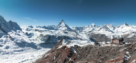 An aerial view of the Matterhorn in Zermatt, Switzerland, shows its pyramid shape against a blue sky. The photo reveals snow covered terrain and a mountain lodge, suggesting a serene alpine setting.
