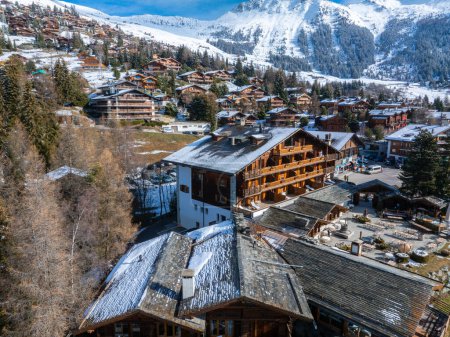 Foto aérea de Verbier, Suiza, muestra edificios de chalets con techos cubiertos de nieve en un paisaje de montaña, capturando la tranquilidad y la belleza del invierno en esta ciudad estación de esquí.