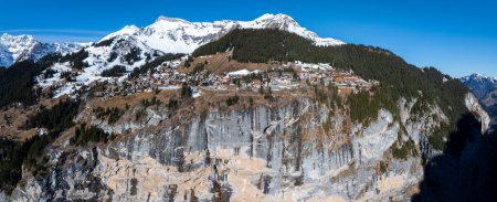 Aerial view of Murren, Switzerland, shows the town on a cliffs edge against snow capped mountains. Traditional alpine buildings and dense forests under a clear blue sky highlight its serene beauty.