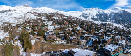 Aerial view of Verbier, Switzerland shows chalet buildings amid snow and greenery. Roads wind under a blue sky with snow capped mountains, marking it a top winter sports spot.