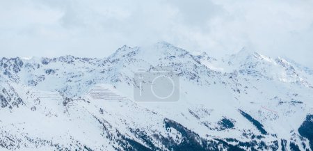 Un plan aérien montre Verbier, un paysage enneigé de Suisse, des pistes de ski, un pic et les Alpes sous un ciel partiellement nuageux. Son charme hivernal est dans la neige intacte et de douces ombres nuageuses.
