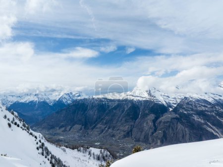 An aerial shot shows Verbier, Switzerlands snowy landscape, ski tracks, a peak, and the Alps under a partly cloudy sky. Its winter charm is in the untouched snow and gentle cloud shadows.