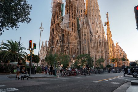 Téléchargez les photos : Barcelonas rue calme à l'heure dorée, la Sagrada Familia tourne derrière. Ciel dégagé, lumière rouge et habitants près d'un passage piétonnier avec des vélos créent une ambiance animée et conviviale. - en image libre de droit