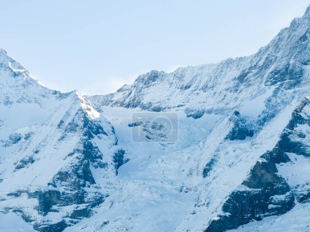 Una toma aérea muestra Verbier, paisajes nevados de Suiza, pistas de esquí, un pico y los Alpes bajo un cielo parcialmente nublado. Su encanto invernal está en la nieve intacta y las sombras suaves de las nubes.