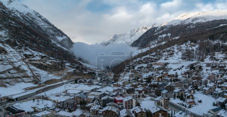 Luftaufnahme von Zermatt, Schweiz, zeigt Chalets und Hotels mit Schneedächern. Der Matterhorngipfel sticht hervor, mit einer Zahnradbahn, die sich durch die Stadt schlängelt, gegen verschneite Hänge und Berge.