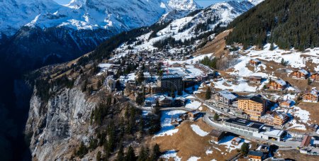 Vista aérea de Murren, Suiza, muestra un sereno pueblo de montaña con edificios de estilo chalet tradicional en un acantilado. Los Alpes suizos cubiertos de nieve y los cielos despejados crean un escenario pintoresco.