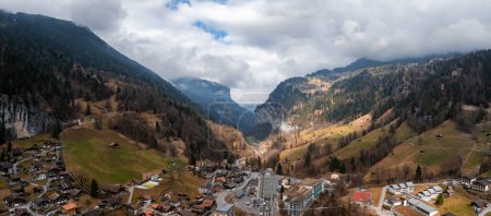 Aerial view of Murren, Switzerland, reveals its serene alpine charm. Chalets with sloping roofs along the main street merge into forested mountains. An overcast sky creates dynamic shadows