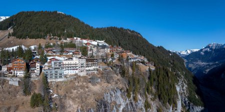 Aerial view of Murren, Switzerland, showcases a serene mountain village with traditional chalet style buildings on a cliff. Snow covered Swiss Alps and clear skies create a picturesque backdrop.