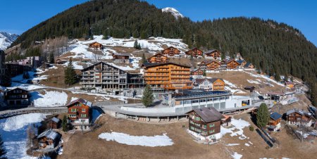 Luftaufnahme von Murren, Schweiz, zeigt ein ruhiges Bergdorf mit traditionellen Gebäuden im Chalet-Stil auf einer Klippe. Schneebedeckte Schweizer Alpen und klarer Himmel schaffen eine malerische Kulisse.