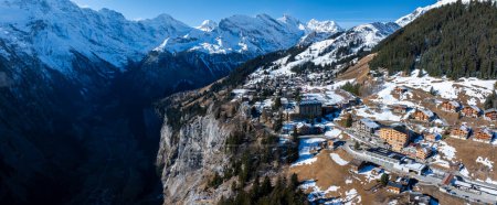 Aerial view of Murren, Switzerland, showcases a serene mountain village with traditional chalet style buildings on a cliff. Snow covered Swiss Alps and clear skies create a picturesque backdrop.
