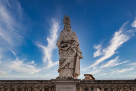 Estatua de piedra de figura religiosa en la Ciudad del Vaticano, adornada con vestimentas eclesiásticas, sosteniendo un libro. Contra el cielo azul con nubes, indicios de grandeza.