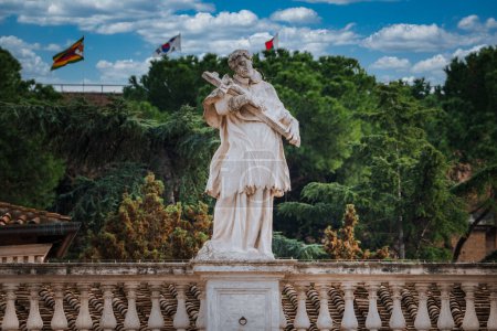 Classical statue of robed figure holding a cross in Vatican, against lush greenery and blue sky. Flags, balustrade, serene atmosphere. Rich cultural heritage showcased.
