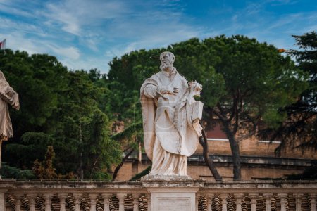 Estatua de mármol de una figura barbuda en la Ciudad del Vaticano, de pie sobre un pedestal con balaustres clásicos. Envejecido debido al envejecimiento, sobre un fondo de exuberantes árboles verdes y parcialmente nublado cielo azul.