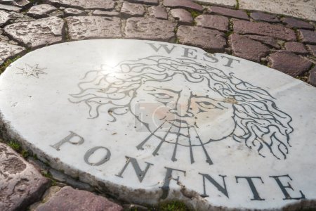 Vista de cerca de una piedra de mármol blanco en la Ciudad del Vaticano con un rostro estilizado e inscripción que representa un dios del viento o figura similar, marcando la dirección occidental.