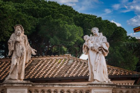 Weathered statues of a bearded figure and a figure holding a child on a Mediterranean tiled roof with lush green trees in the background. Likely in the Vatican.