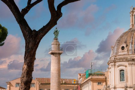 Historic European city scene featuring a large dome, ancient victory column with statue, leafy tree, and cloudy sky. Likely Rome near the Roman Forum.