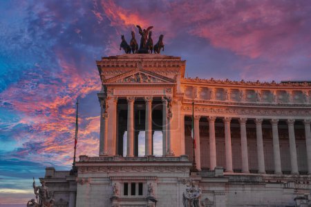 Neoclassical building at sunrise or sunset in Italy. Tall columns, pediment with sculptures, and a bronze chariot sculpture on top. Flag with green, white, and red stripes. Latin inscriptions.