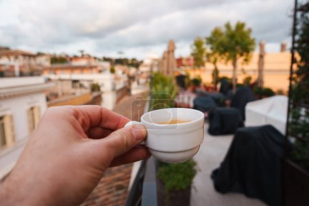 Terraza de lujo en Roma primer plano de la mano sosteniendo taza de café blanco, techos de edificios de colores cálidos en el fondo, toques de vegetación y cielo nublado. Atmósfera relajante.
