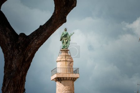 Foto de Estatua de bronce de figura clásica sobre columna de piedra con plataforma de observación, rodeada de barandilla. Cielo nublado con rama de árbol en primer plano, ubicación desconocida. - Imagen libre de derechos