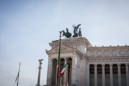 Neoclassical building in Rome with grand columns, bronze sculptures, and Italian flags fluttering under a clear sky. Iconic monument honoring Victor Emmanuel II.