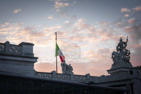 Ein friedlicher Himmel in rosa und orangefarbenen Tönen mit einer italienischen Flagge, die sanft im Wind flattert. Ornamentale Skulpturen und architektonische Details deuten auf einen bedeutenden historischen Ort in Italien hin.