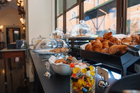 Breakfast buffet in a luxury hotel in Rome elegant setup with pastries, bread rolls, croissants, and candies. Urban view visible through large windows. Luxurious ambiance with modern decor.