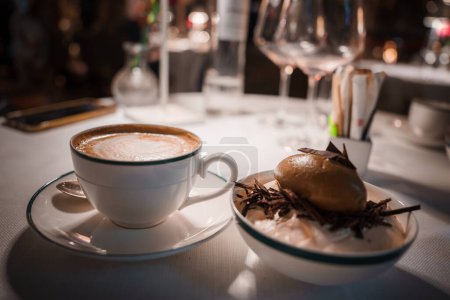 Close up view of elegant table setting in a high end restaurant. White cup of cappuccino and chocolate dessert on a white tablecloth with dining utensils. Rich, warm ambiance.