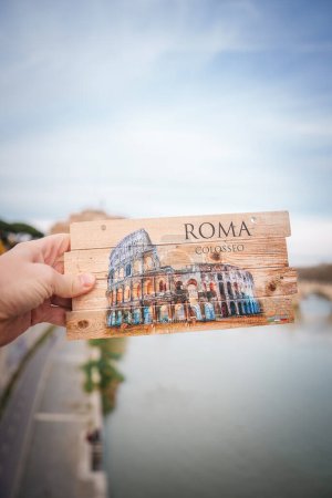 Téléchargez les photos : Main tenant une carte postale en bois avec une illustration du Colisée de Rome, Italie. Affichage ROMA et COLOSSEO, design rustique sur fond de bois près de la rivière et du pont. - en image libre de droit