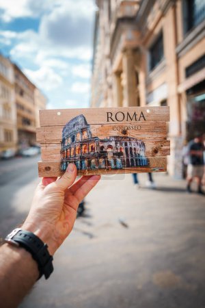 Photo for Hand holding souvenir with Colosseum depiction, ROMA and COLOSSEO printed on wood. Background of Rome street with people, European buildings. Sky partly cloudy. Main focus is on souvenir. - Royalty Free Image